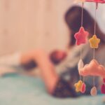 A mother cradling her baby next to a dreamy cloud and star mobile in a soft-focus bedroom setting.